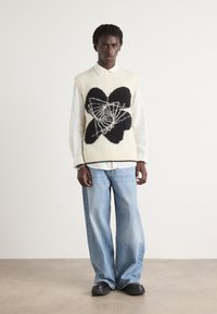 Suéter sin mangas de punto en crema con un gran diseño floral negro y acentos blancos, llevado sobre una camisa blanca con cuello y combinado con vaqueros de pierna ancha azul claro.