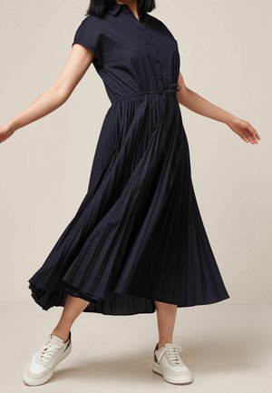Robe midi plissée marine à manches courtes, fermeture à boutons sur le devant et taille à cordon. Associée à des baskets blanches.
