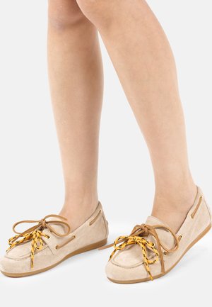 Chaussures mocassin en daim beige avec des lacets à motifs marron et jaune portées pieds nus sur un fond uni.