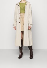 Beige langer Trenchcoat mit breitem Kragen, braunen Knöpfen und seitlichen Taschen, drapiert über einem grünen Oberteil und einem beigen Rock, kombiniert mit braunen Stiefeln.