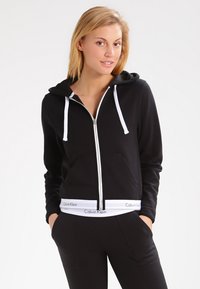 Sort zip-hoodie med hvide snøre og elastik i taljen. Har frontlommer og blødt stof, designet til komfort og afslappet brug.