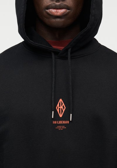 Juodas džemperis su gobtuvu, pagamintas iš minkštos medžiagos, su raudonu logotipu ir tekstu ant krūtinės, su raišteliais ir metalinėmis galvutėmis. Paprastas dizainas.