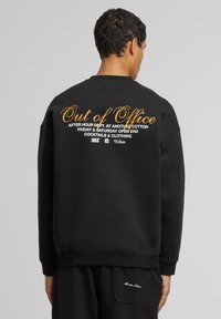 Sudadera de algodón negra con texto gráfico en blanco y naranja en la parte trasera. El diseño incluye un corte casual con puños y dobladillo acanalados.