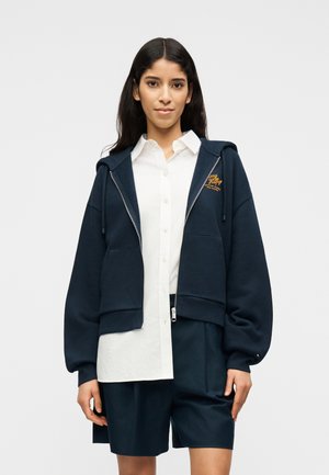 Γυναίκα με μπλε φούτερ με φερμουάρ πάνω από λευκό πουκάμισο και μπλε σορτς, στέκεται μπροστά σε απλό ανοιχτό φόντο.