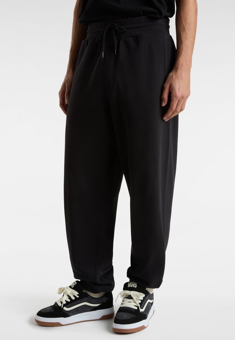 Černé tepláky s páskem na stažení v pase, zúženým střihem a žebrovanými manžetami u kotníků, doplněné černobílými teniskami s kontrastním logem.