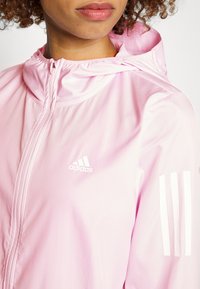 Jasnoróżowa kurtka Adidas z zamkiem błyskawicznym, z teksturowaną powierzchnią, kapturem i białymi trzema paskami na rękawie, logo na piersi.