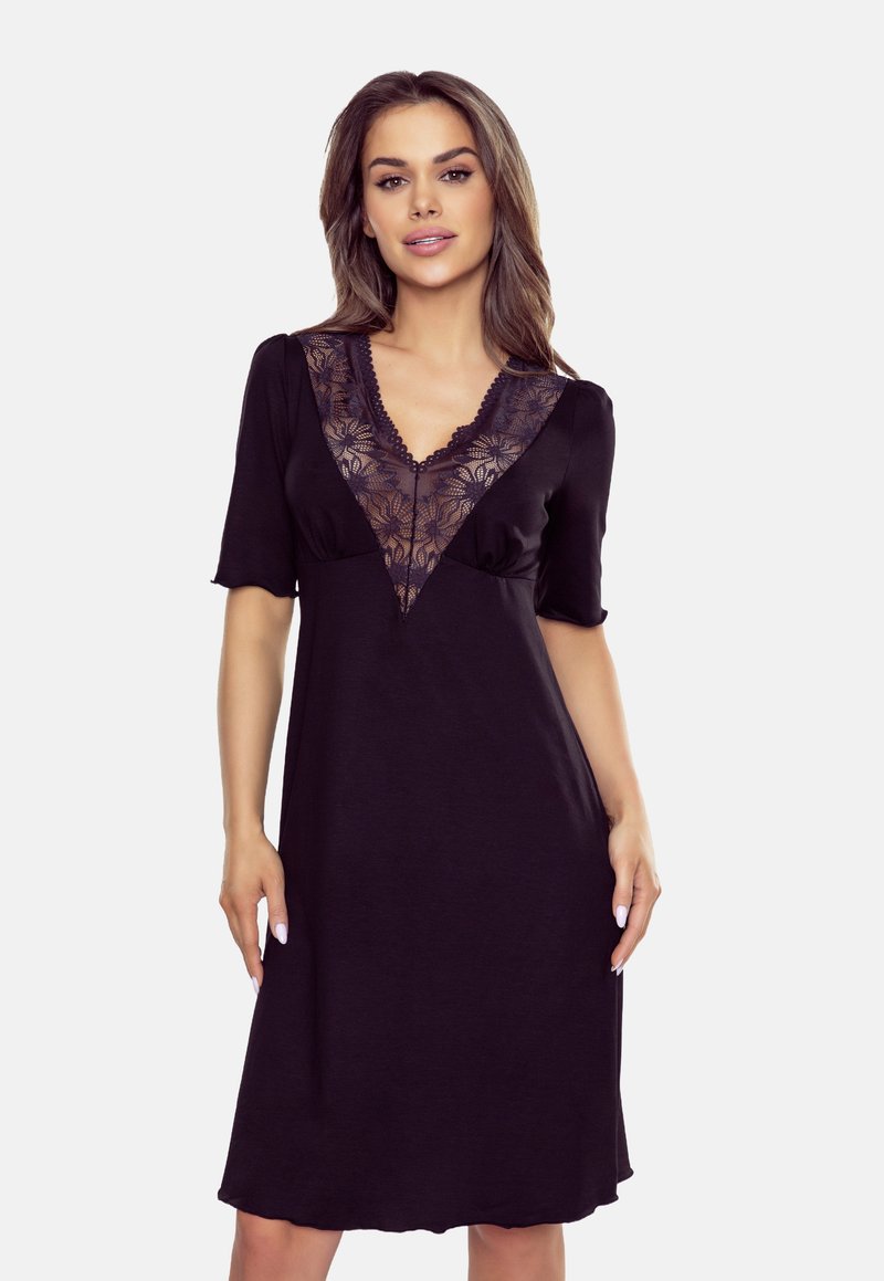 Modelka pozuje w czarnej sukience z przezroczystym koronkowym dekoltem w kształcie litery V i krótkimi rękawami, na jasnoszarym tle. Jej włosy są stylizowane, a makijaż schludny.