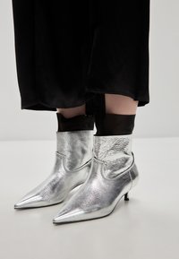 Botines metálicos plateados con punta afilada, con un diseño recortado en la parte superior y un tacón kitten bajo, combinados con tejido negro.