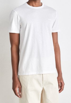 Muž v jednobarevném bílém tričku s krátkým rukávem a světle béžových kalhotách na jednobarevném pozadí.