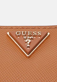 Surface en cuir marron texturée avec un logo triangulaire doré Guess USA comportant un point d'interrogation au centre.