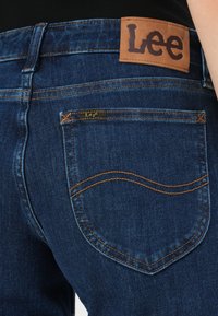 Sötétkék farmernadrág bőr címkével, amelyen az "Lee" felirat olvasható a derékszíjon. Kontrasztos narancssárga varrás jellemzi, valamint két hátsó zseb található.