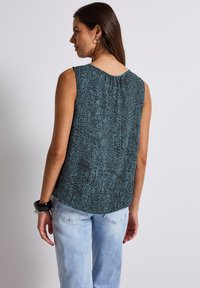 Sleeveless teal top with a textured crocodile pattern, rounded neckline, and a pleated detail at the back, paired with light jeans.