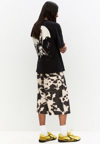 Maglietta grafica nera con un grande design sulla schiena, abbinata a pantaloni shorts a stampa mucca di lunghezza fino al ginocchio e sneakers gialle. Calzini bianchi visibili.