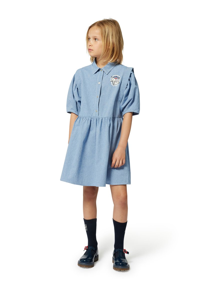 Robe en jean bleu clair avec des manches bouffantes courtes, un devant à boutons et une jupe froncée. Elle dispose d'un patch brodé sur la poitrine.