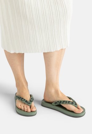 Füße, die grüne Flip-Flops mit schwarzen Riemen mit Tiermuster tragen, kombiniert mit einem weißen gerippten Midirock vor einem einfarbigen hellen Hintergrund.
