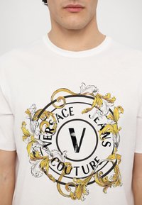 Balta medvilninė marškinėliai su apskritimu juodos ir auksinės spalvos dekoratyviniu grafišku dizainu, kuriame užrašyta "VERSACE JEANS COUTURE" ir stilizuotas "V" centre.