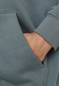 Grijze sweatshirt met een voorzak waarin een hand gedeeltelijk zichtbaar is. De stof is zacht en glad, met subtiel gestikte naden.