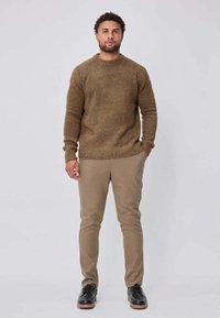 Homme debout, portant un pull en maille marron, un pantalon beige et des chaussures noires, avec une main dans la poche, sur un fond clair uni.