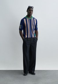 Άνδρας στέκεται μπροστά σε απλή επιφάνεια τοίχου, φοράει μπλε και μπεζ ριγέ πουκάμισο, μαύρο παντελόνι με φαρδύ πόδι, μαύρα παπούτσια και γυαλιά ηλίου στο κεφάλι.