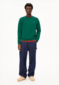 Felpa verde con colletto rosso, decorata con un piccolo logo bianco; abbinata a jeans blu larghi e pantofole beige.