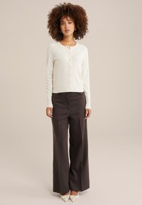 Ivory džemperis ar apaļu kakla izgriezumu un pogu priekšpusi, apvienots ar plaša griezuma tumšām biksēm un smailām baltām kurpēm.