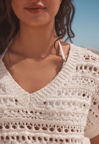 Witte gehaakte top met V-hals, openweefpatroon en korte mouwen, die een witte bikini eronder onthult.