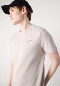 Camiseta de color beige claro con mangas cortas, escote redondo y el logo de Calvin Klein en letras oscuras en el lado izquierdo del pecho. Textura suave.