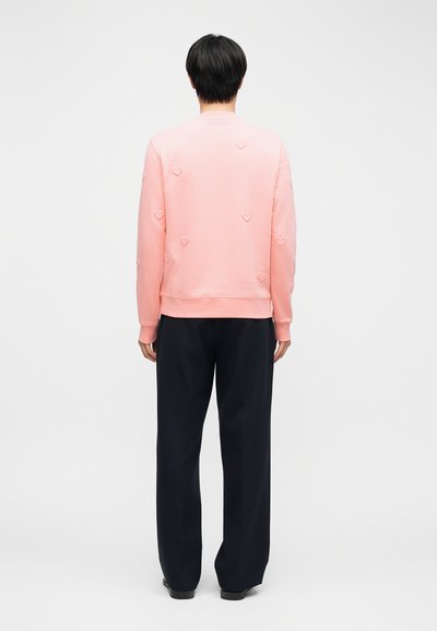 Άτομο που στέκεται με την πλάτη γυρισμένη, φορώντας ροζ φούτερ με ανεβασμένες καρδιές και μαύρο παντελόνι φαρδύ, μπροστά σε ένα απλό φόντο.