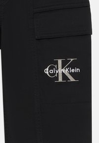 Tela negra con grandes iniciales "cK" cosidas y texto "Calvin Klein" bordado en blanco en una solapa de bolsillo.
