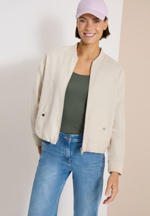 Smilende person iført beige lynlåstrøje, grøn top, blå jeans og lyserød kasket, der står mod en to-farvet beige baggrund.