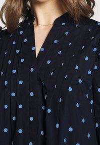 Marinblus med blåa prickar, med V-ringning, veckad design och volangdetaljer på ärmarna. Lätt, texturerad tyg.