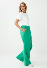 Witte korte mouwen shirt gecombineerd met losse, felgroene, elastische broek met taille. Het model staat met één hand in de zak en draagt witte sneakers.