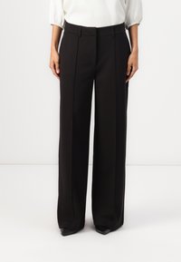 Pantalon noir à jambes larges avec une texture lisse, doté de plis à l'avant et d'une coupe ajustée, associé à des chaussures noires pointues.
