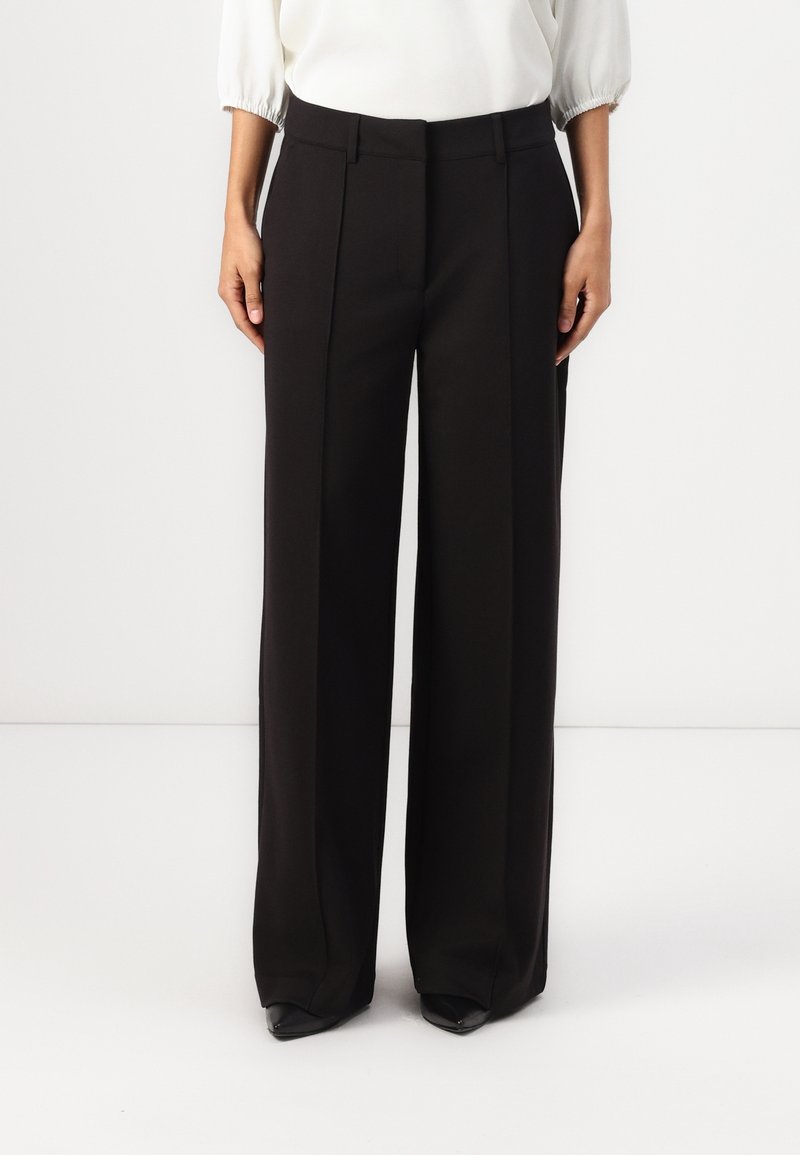 Pantalon noir à jambes larges avec une texture lisse, doté de plis à l'avant et d'une coupe ajustée, associé à des chaussures noires pointues.
