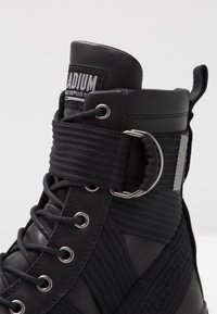 Černá vysoká botka s texturovaným svrškem, textilními popruhy, kovovým příslušenstvím a logem. Šnurování s kovovými očky pro uzavření.