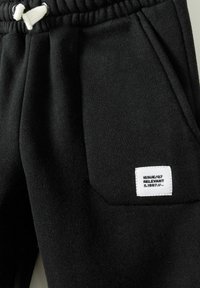 Zwarte joggingbroek van een gestructureerde stof, met een elastische tailleband, een witte trekkoord en een klein wit vierkant label op de zak.