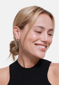 Boucles d'oreilles en or en forme de hoop avec une surface texturée, comportant de petites pierres claires. Le design est circulaire avec un fermoir minimaliste.