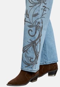 Vaqueros de denim de color azul claro con intrincados diseños florales bordados en marrón a lo largo del lado exterior, combinados con botas de tobillo de ante marrón oscuro.