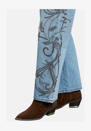 Jeans di denim azzurro chiaro con intricate decorazioni floreali ricamate in marrone lungo il lato esterno, abbinati a stivaletti alla caviglia in suede marrone scuro.
