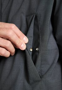 Dunkelgrauer Stoff, der eine Hand zeigt, die die Tasche eines Kleidungsstücks greift, mit einem Druckknopfdetail und einer sauberen, strukturierten Oberfläche.