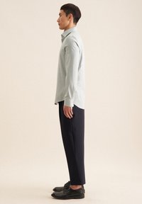 Camicia verde chiaro a maniche lunghe con colletto, abbinata a pantaloni scuri e scarpe nere. La camicia presenta un taglio dritto e spacchetti laterali.