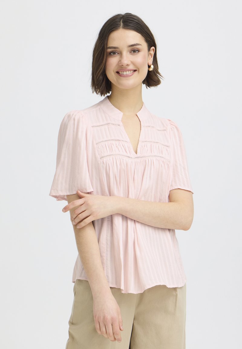 Jonge vrouw met kort donker haar, gekleed in een lichtroze blouse en beige broek, glimlachend met zachtjes gekruiste armen.