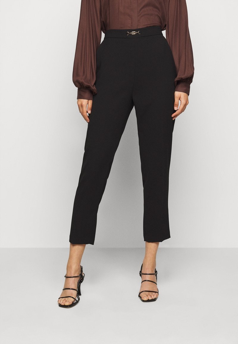Femme portant un pantalon court noir avec une ceinture décorative, un chemisier marron à manches longues et des sandales à talons noires avec des lanières.