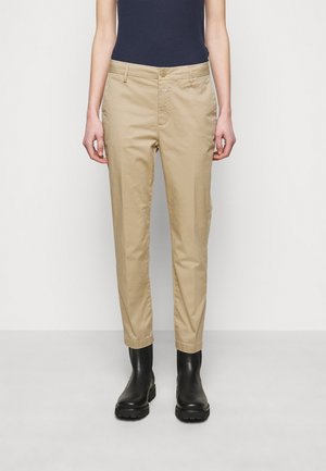 Pantalon beige slim à bouton et fermeture éclair devant, porté avec des bottines noires et un haut sans manches foncé, sur un fond blanc uni.