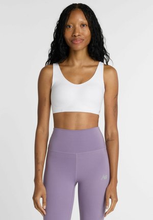 Donna con lunghi capelli neri e ricci che indossa un reggiseno sportivo bianco e leggings lavanda a vita alta su uno sfondo chiaro uniforme.