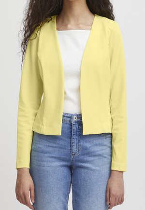 Frau trägt ein hellgelbes, vorne offenes Sakko über einem weißen Top und blauen Jeans, steht vor einem einfarbigen Hintergrund.