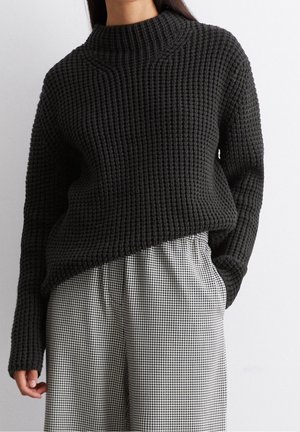 Femme portant un pull en grosse maille noire rentré dans un pantalon large taille haute à carreaux noir et blanc, mains détendues.