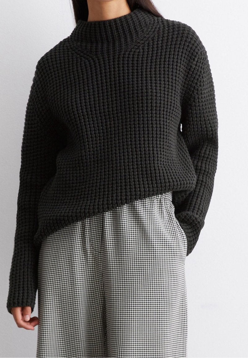 Femme portant un pull en grosse maille noire rentré dans un pantalon large taille haute à carreaux noir et blanc, mains détendues.