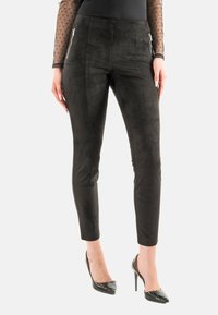 Pantalones de ante sintético negros con corte ajustado, que cuentan con bolsillos con cremallera en la parte delantera y una textura suave y mate. Combinados con tacones altos negros.