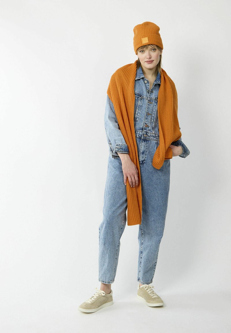 Denim overalls in light blue, paired with a ribbed orange scarf and beanie. Beige sneakers complete the look, featuring a simple, modern design.